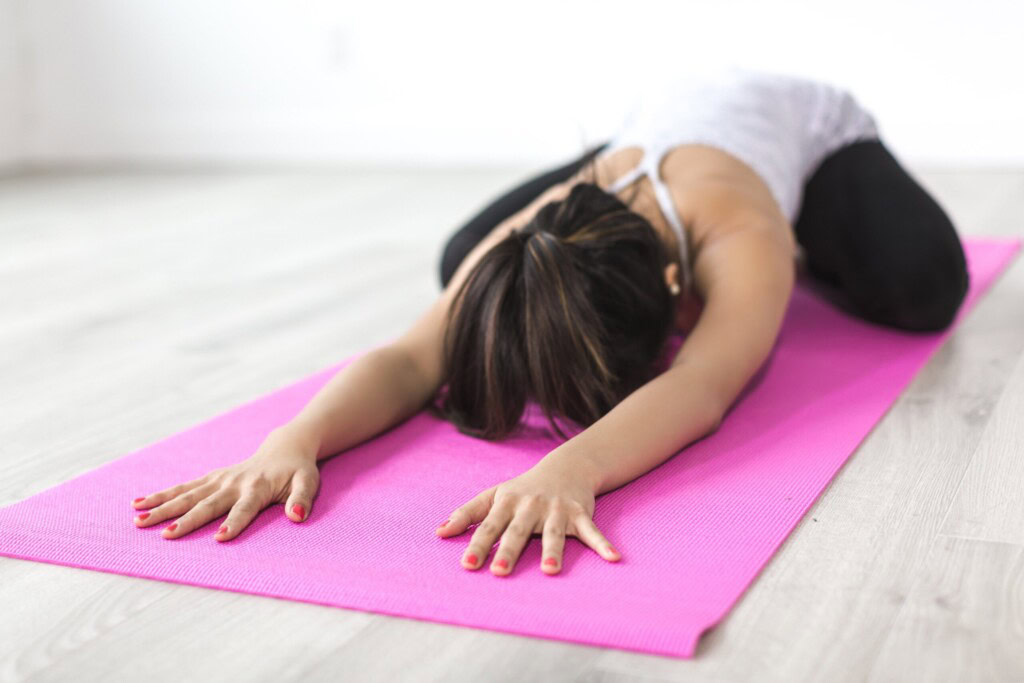Yoga practitioner performing Ardha-Kurmasana (Half Tortoise Pose), kneeling on the mat and extending the arms forward, lowering the chest toward the floor, while the forehead reaches towards the ground. The pose stretches the back, shoulders, and arms, while promoting deep relaxation and flexibility