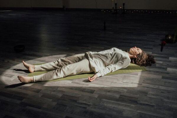 Yoga practitioner lying on their back in Savasana (Corpse Pose), with arms relaxed at the sides and legs extended comfortably apart. The pose promotes deep relaxation, calms the mind, and allows the body to absorb the benefits of the practice